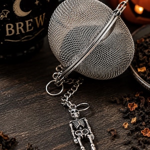 Puede incluir: Un infusor de té esférico de acero inoxidable con una malla fina y cierre de bisagra. Una cadena de metal plateado está unida al infusor y termina en un dije de esqueleto detallado que cuelga de un pequeño anillo circular.