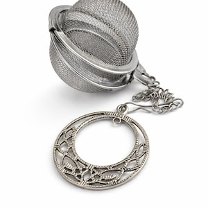 Może przedstawiać: Stalowy zaparzacz do herbaty z siatkową kulą i ozdobnym, ażurowym pierścieniem. Zaparzacz przeznaczony jest do przechowywania liści herbaty i jest przymocowany do łańcuszka.