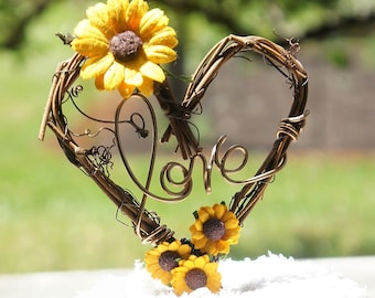 May include: A rustic wedding cake topper featuring a heart-shaped wire frame with the word "love" inscribed. The frame is decorated with small yellow sunflowers.