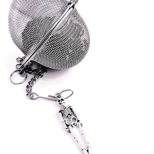 Puede incluir: Un infusor de té de plata con forma de esfera, con un asa y una cadena unida a un amuleto de esqueleto.