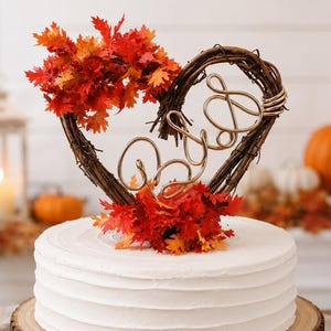 May include: A cake topper featuring a heart-shaped twig frame adorned with vibrant red and orange autumn maple leaves. Centered within the frame is a gold-toned wire script reading "B&D". The topper sits atop a plain white frosted cake.