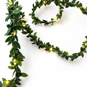 Puede incluir: Una guirnalda decorativa con hojas artificiales de color verde oscuro entrelazadas con hilos de alambre finos y flexibles. Pequeñas luces LED de tonos cálidos están espaciadas uniformemente por el follaje, creando un efecto brillante.