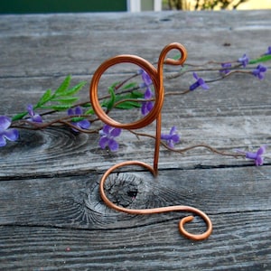 Può includere: Una lettera 'G' in filo di rame a forma di supporto decorativo. Il supporto è su una superficie di legno con fiori viola.