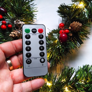 Puede incluir: Un mando a distancia blanco con un botón verde "ON", un botón rojo "OFF" y un botón gris "TIMER". El mando tiene botones numerados del 1 al 8, un botón "MODE" y un botón "DIM" con signos más y menos. El mando se sostiene en una mano contra un fondo de una guirnalda navideña verde con bayas rojas y luces blancas.