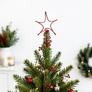 May include: A red wire star tree topper featuring a coiled base, positioned atop a green pine Christmas tree decorated with small red and silver spherical ornaments and clusters of red berries, creating a festive holiday display.