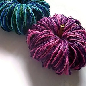 May include: Two yarn pumpkins, one blue and green, the other pink and purple. Both pumpkins have a twig stem.