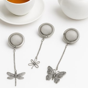 Könnte beinhalten: Drei silberne Tee-Siebe mit dekorativen Anhängern, darunter eine Libelle, eine Blume und ein Schmetterling. Eine weiße Teekanne und eine Teetasse mit Tee befinden sich im Hintergrund. Die Siebe sind aus Metall und haben einen Netzkorb.