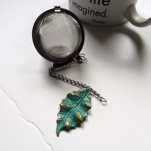 May include: A silver tea infuser with a chain and a green leaf charm. The infuser is round and has a fine mesh. The leaf charm is detailed and has a rustic finish.