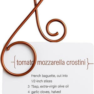 May include: A copper-colored recipe holder with a white card featuring the text "tomato mozzarella crostini" and a list of ingredients. The holder has a decorative, looping design.