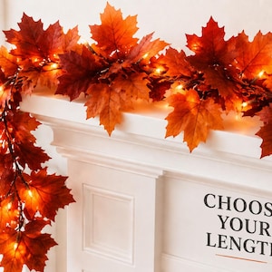 May include: A decorative garland featuring artificial maple leaves in shades of orange, red, and yellow, interspersed with small warm white LED lights. The garland is draped over a white mantelpiece with the text CHOOSE YOUR LENGTH printed on it.