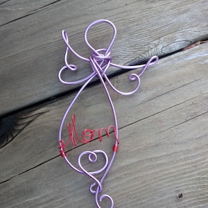 Peut inclure: Une sculpture en fil de fer décorative en forme d'ange avec du fil couleur lavande formant la tête, les ailes et le corps. Le mot Mom est écrit en fil rouge au centre du corps de l'ange, et un petit cœur est formé à la base.