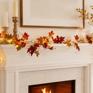 May include: An artificial autumn garland featuring realistic maple leaves in shades of red, orange, and yellow, intertwined with small warm white LED string lights. The flexible vine drapes across a white mantelpiece, creating a seasonal display.