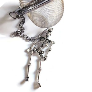 May include: A silver tea infuser shaped like a sphere with a skeleton hanging from the chain. The infuser has a mesh design and a hinged lid.
