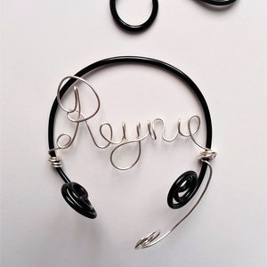 May include: A silver wire bracelet with the name "Rayne" spelled out in wire. The bracelet is attached to a black wire hoop with two black wire loops on the sides.
