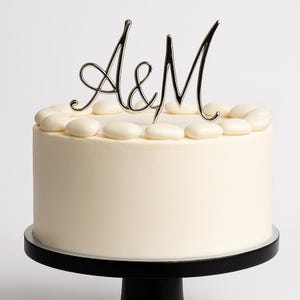 Może przedstawiać: Okrągłe, białe ciasto z srebrnym topperem na ciasto "A&M". Ciasto jest udekorowane białymi, owalnymi cukierkami na górnej krawędzi. Ciasto stoi na czarnym stojaku na ciasto.
