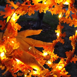 May include: A string of warm white fairy lights wrapped around a garland of orange and brown autumn leaves. The lights are lit and create a warm glow.