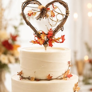 May include: A two-tier white frosted cake topped with a heart-shaped twig topper featuring gold wire letters J&D and small orange, red, and yellow autumn leaves. A thin vine with matching fall foliage wraps around the base of the top cake tier.