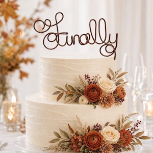 May include: A two-tier white frosted cake topped with a dark brown wire cake topper featuring the word Finally in cursive script. The cake is decorated with clusters of orange, cream, and brown flowers, dried leaves, and small berries.