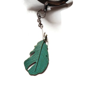Może przedstawiać: Metalowy, turkusowy zawieszek w kształcie pióra przymocowany do srebrnego łańcuszka. Łańcuszek jest połączony z metalowym zaparzaczem do herbaty.