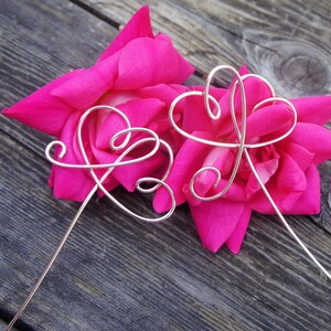 Può includere: Due forme di cuore in filo d'argento sono intrecciate e posizionate su due petali di rosa rosa. I cuori di filo sono su una superficie di legno.