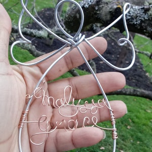 May include: A handcrafted wire art piece in silver and copper tones. The design features a stylized angel shape with the names "Malyssa Grace" written in cursive. The piece is held in a hand against a blurred outdoor background.