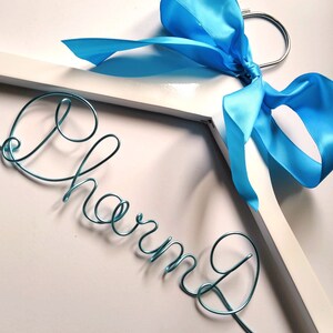 May include: A white wooden hanger with a blue satin ribbon tied in a bow. The word "Charm" is spelled out in turquoise wire.