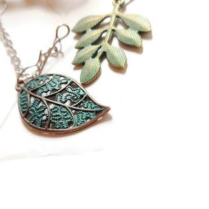 May include: Close-up of two tea infusers with leaf-shaped charms. One charm is a detailed leaf design in teal and bronze, the other is a sprig of leaves in a similar color. The infusers are made of a fine mesh.