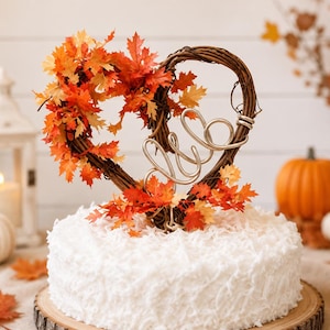 May include: A cake topper featuring a heart-shaped brown twig wreath adorned with orange, red, and yellow artificial maple leaves, centered with silver wire cursive letters, placed atop a round white coconut-covered cake on a wooden base.