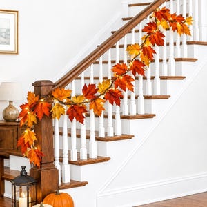 May include: An artificial autumn garland featuring vibrant maple leaves in shades of red, orange, and yellow, intertwined with small warm white LED lights. The garland is draped along a wooden staircase railing, creating a seasonal decorative display.