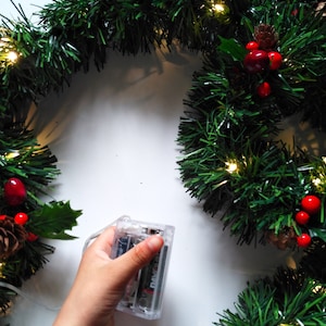 Op de afbeelding: Een hand houdt een doorzichtig plastic batterijpakket vast voor een kerstverlichtingssnoer. De lampjes zijn om een ​​groene krans gewikkeld met rode bessen en dennenappels.