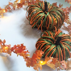 Op de afbeelding: Twee kleine pompoenen gemaakt van touw, een groen en oranje, de andere oranje en groen, zijn omringd door een krans van oranje bladeren met kleine witte lampjes.