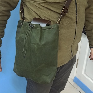 May include: Olive green canvas shoulder bag with a drawstring closure. The bag has a brown leather flap and a brown leather shoulder strap with brass-colored hardware. The bag is worn over the shoulder.