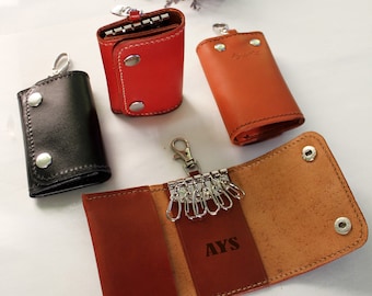 May include: A collection of leather key holders in various colors: black, red, and orange. One key holder is open, displaying key rings and the initials "AYS". The key holders have snap closures and metal hardware.