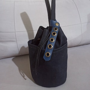 May include: A black canvas bucket bag with a blue leather strap and gold metal eyelets. The bag has a drawstring closure and a single top handle.