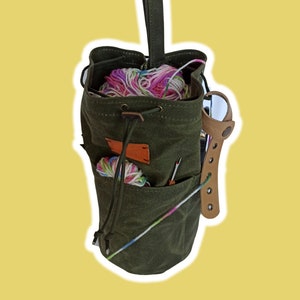 Può includere: Una borsa per filati in tela verde con chiusura a coulisse e cinturino in pelle. La borsa è piena di filati colorati e ferri da maglia.