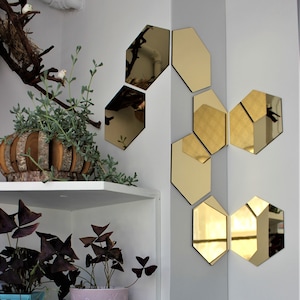 May include: A set of six gold hexagonal mirrors arranged in a corner of a white wall. The mirrors reflect the surrounding room, including a shelf with plants and a white shelf.
