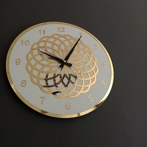 Puede incluir: Una esfera de reloj blanca con marcadores de hora romanos dorados y un dise&ntilde;o geom&eacute;trico dorado. Las manecillas del reloj son negras.