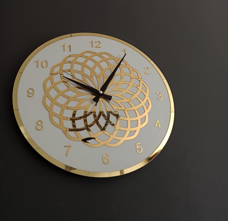 Puede incluir: Una esfera de reloj blanca con marcadores de hora romanos dorados y un dise&ntilde;o geom&eacute;trico dorado. Las manecillas del reloj son negras.