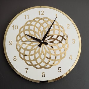 Puede incluir: Un reloj redondo blanco con n&uacute;meros romanos dorados y un dise&ntilde;o geom&eacute;trico dorado en el centro. El reloj tiene agujas negras.