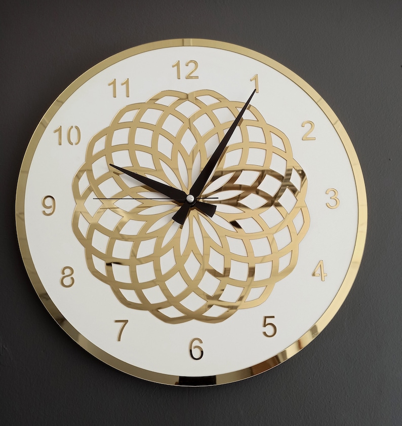 Puede incluir: Un reloj redondo blanco con n&uacute;meros romanos dorados y un dise&ntilde;o geom&eacute;trico dorado en el centro. El reloj tiene agujas negras.