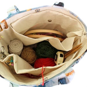 May include: A fabric tote bag with a blue patterned exterior and a cream-colored interior featuring multiple pockets. The bag contains several balls of yarn in red, green, and brown, a wooden embroidery hoop, knitting needles, and scissors.