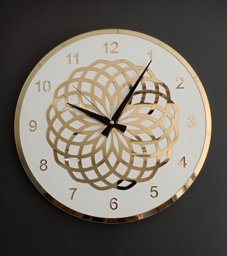 Puede incluir: Una esfera de reloj blanca con marcadores de hora romanos dorados y un dise&ntilde;o geom&eacute;trico dorado en el centro. El reloj tiene agujas negras y un borde dorado.