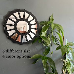 May include: A black circular mirror with a sunburst design. The mirror is hanging on a gray wall. The text "6 different size 4 color options" is below the mirror.