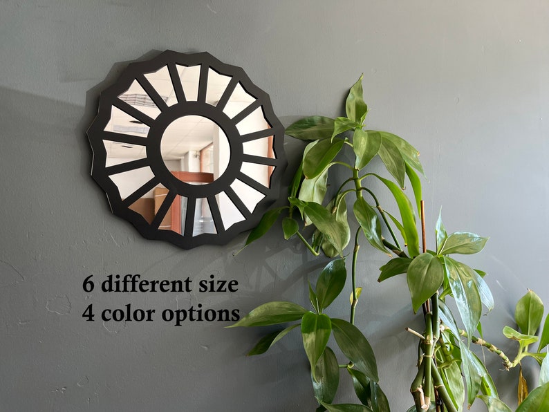 May include: A black circular mirror with a sunburst design. The mirror is hanging on a gray wall. The text "6 different size 4 color options" is below the mirror.