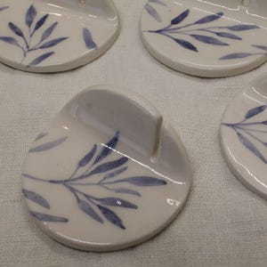 Può includere: Un set di poggiacucchiaio in ceramica bianca con un motivo a foglie blu. Ogni pezzo ha una forma curva con un bordo rialzato. Disposti su una superficie di lino bianco.