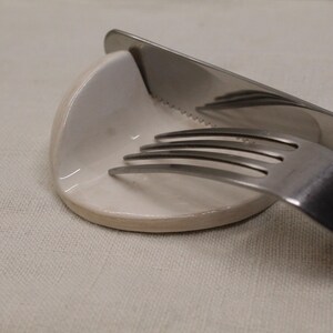 Può includere: Una forchetta e un coltello in acciaio inossidabile poggiano su un piccolo piatto in ceramica bianco sporco. Il coltello è seghettato e angolato sul piatto, con la forchetta appoggiata sulla superficie. Gli oggetti sono su un tessuto di lino chiaro.