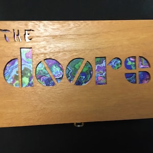 Może przedstawiać: Drewniana skrzynka z wyciętymi słowami "THE DOORS". Litery są wypełnione kolorowym, abstrakcyjnym wzorem w odcieniach fioletu, błękitu i zieleni.