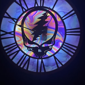 May include: A round wall clock with a black metal frame and Roman numerals. The clock face features a colorful, psychedelic design with a skull and lightning bolt graphic. The hands are black.