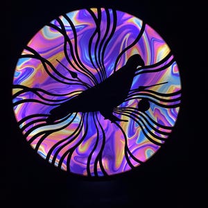Puede incluir: Una obra de arte circular iluminada con la silueta negra de un pájaro sobre un fondo arremolinado de colores morados, azules, amarillos y rosas. Líneas negras curvas enmarcan el pájaro, creando un efecto de capas. La pieza está sobre un fondo negro.