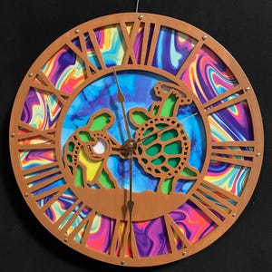 May include: Round wooden wall clock with Roman numerals and a colorful, swirling background. The clock features a laser-cut design of two turtles and gold-colored clock hands. The clock is hanging from a metal wire.